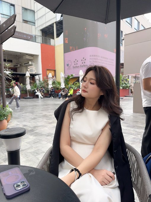 A woman in a white dress sits at an outdoor cafe with a phone on the table, with a 'Forbes Forbes' sign visible in the background.