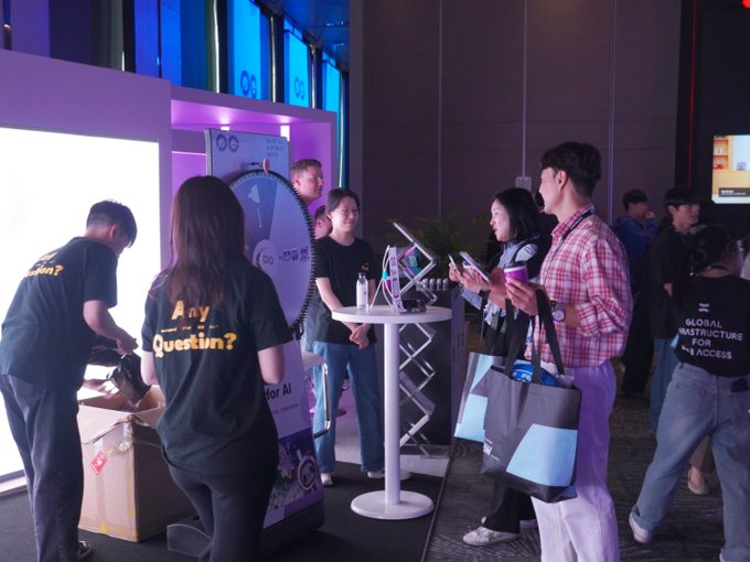 A trade show or event space features people gathered around booths; staff members wear black shirts reading 'Any Question?' and 'GLOBAL INFRASTRUCTURE FOR AI ACCESS'.