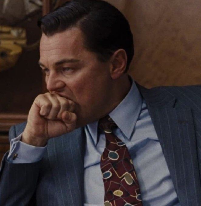 A man in a pinstripe suit and patterned tie rests his hand against his mouth in a contemplative pose.