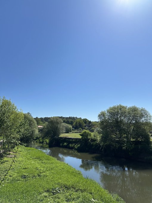 A scenic landscape photo captures a calm river winding through lush green fields and trees under a bright, clear blue sky.