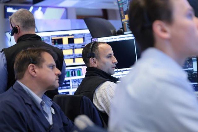 Traders wearing headsets work at a busy trading desk, surrounded by multiple monitors displaying financial charts and data.