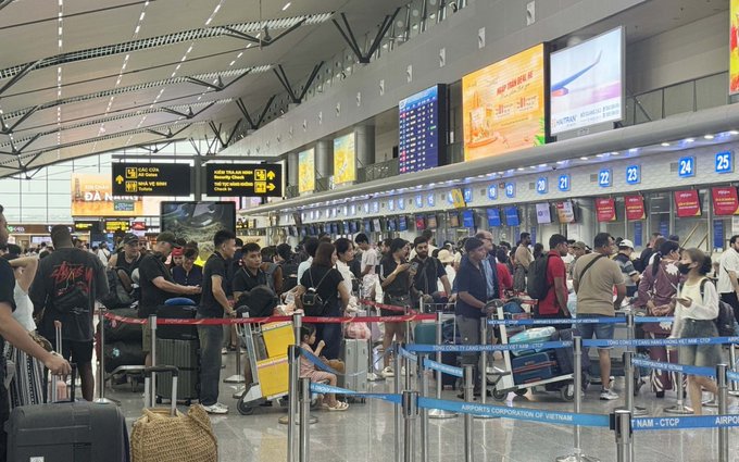 A crowded airport terminal shows travelers with luggage waiting in long check-in lines behind red and blue barriers, with flight information screens and signs for check-in desks (A, B, C, D) and gates (21-25) visible above.
