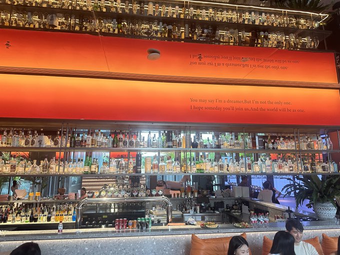 The image shows a bar with many bottles of liquor displayed behind a glass shelf, along with other items such as cups and bowls. The scene takes place at an outdoor restaurant or bar area where people are sitting and enjoying their time. A person is sitting on one side of the image while another individual can be seen standing in front of the bar. There is also a potted plant visible near the center of the scene, adding to the ambiance of the location. The post includes a quote that reads "It's a good idea to have someone you love around when you're dying to go somewhere." This message seems to resonate with the atmosphere and setting of the bar or restaurant where people are gathered.