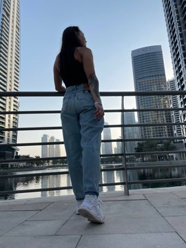 A photo taken from behind of a woman with long dark hair wearing a black crop top and light blue jeans, standing on a balcony overlooking a cityscape with tall skyscrapers.