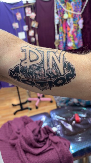 A freshly inked tattoo on a forearm spells out "DTN" above "MOTION," mirroring the post's reference to perpetual motion and the Motion community, with the blurred background showing a tattoo parlor setting complete with clothing racks and a work station.