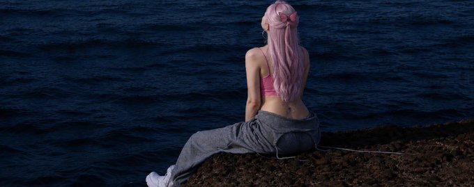 A person with long pink hair sits on a rocky shoreline facing the dark blue sea, wearing a pink top, loose gray pants, and white shoes.