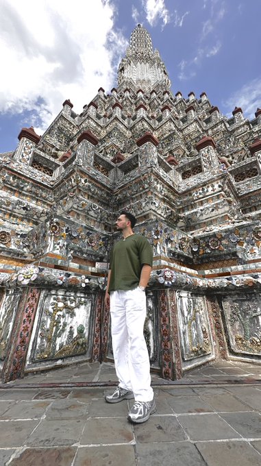 A man in a green shirt and white pants stands posing in front of an ornately decorated temple with intricate tilework and a tall spire.