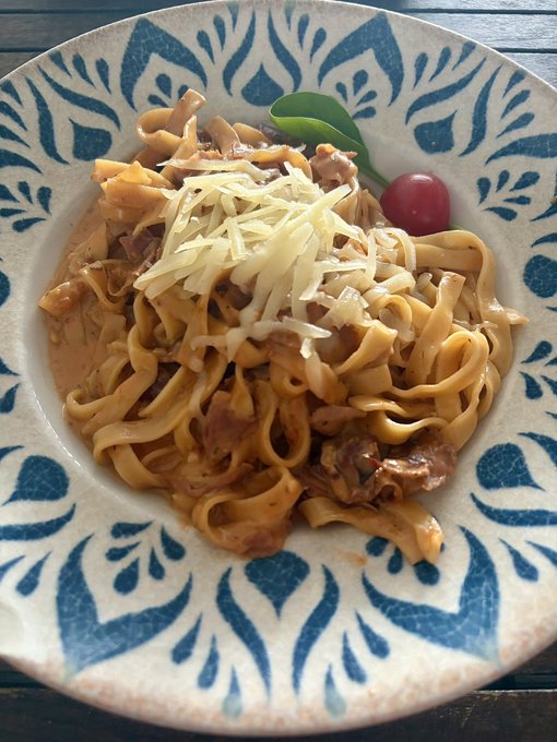 The image shows a plate of pasta with cheese, tomato, and spinach garnish sitting on top of a wooden table. The pasta appears to be a type of spaghetti dish, possibly fettuccine or lasagna noodles. There are also some tomatoes present in the dish. The background is blurred, but it seems like there might be more food items or dishes around the plate.
