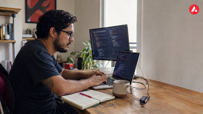 A man wearing glasses sits at a wooden desk in front of a dual-monitor setup with code displayed, accompanied by a laptop, notebook, coffee mug, and houseplants.