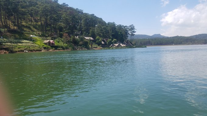 A scenic view of a calm green lake surrounded by pine-forested hills with small wooden cabins along the shoreline under a partly cloudy sky.