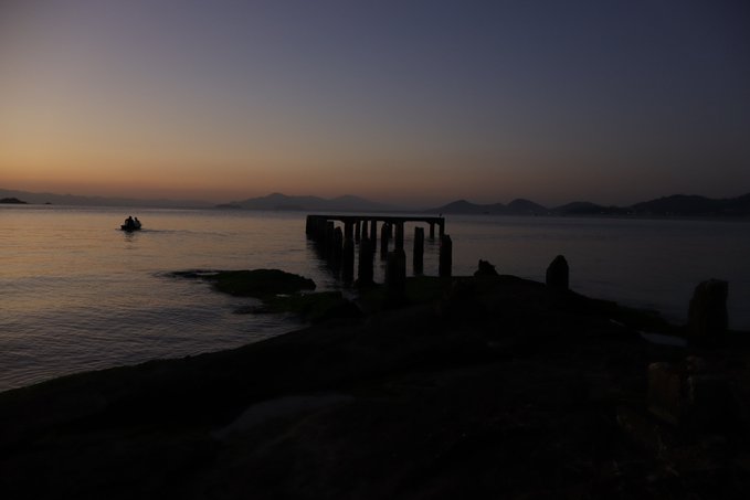 A serene twilight seascape featuring silhouettes of an old pier and a small boat on calm water with distant mountains on the horizon.