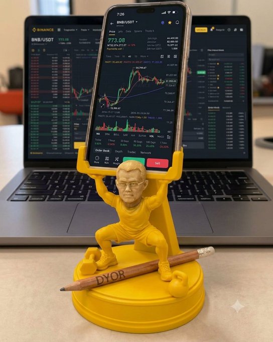 A yellow figurine of a man holding up a phone displaying a Binance (BNB/USDT) trading chart at $773.08, standing on a pencil engraved with 'DYOR', with a laptop showing similar charts in the background.