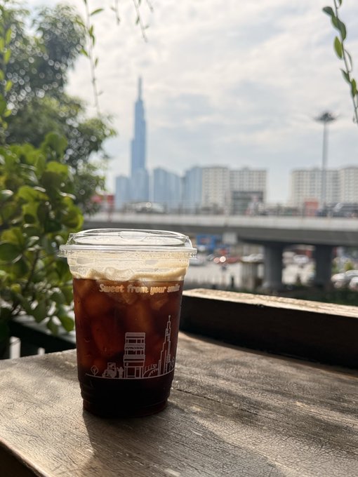 A plastic cup of iced coffee sits on a wooden ledge with a city skyline, including a tall skyscraper, visible in the background.