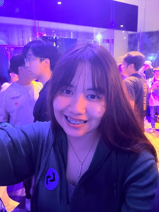 A young woman with braces smiles while taking a selfie in a purple-lit venue, wearing a dark jacket with a logo pin and a necklace.