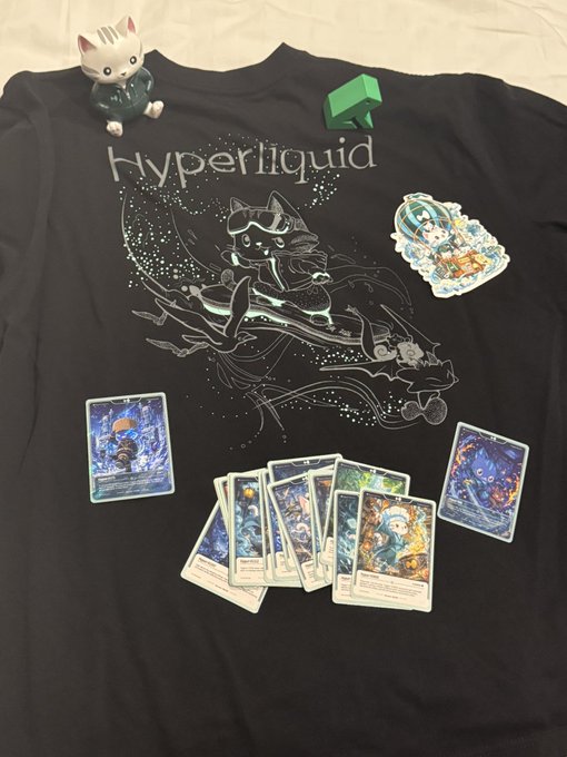 A Hyperliquid-branded black t-shirt is displayed alongside a maneki-neko figurine, a green cash stack toy, and a fan of trading card game cards—visually echoing the post's playful tone about the Hyperliquid team despite past teasing.