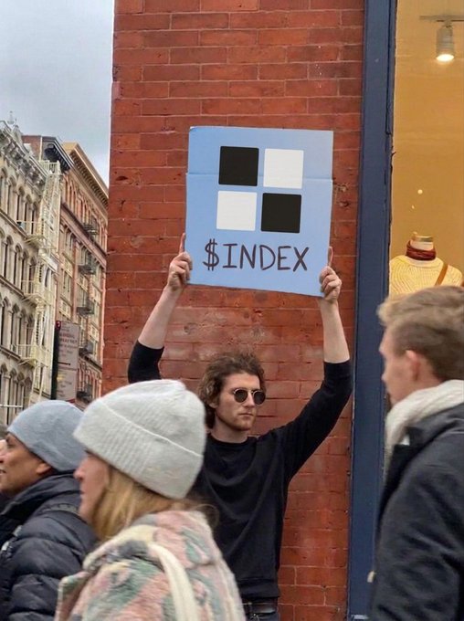 A man wearing sunglasses stands on a city street holding up a light blue protest sign featuring a four-square logo and the text '$INDEX'.