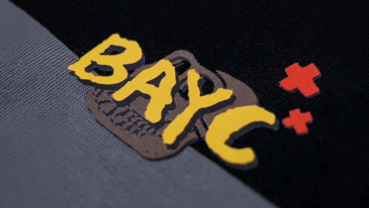 A close-up of fabric shows yellow graffiti-style text reading 'BAYC' layered over a brown ape skull design, accompanied by two red 'x' marks on black material.