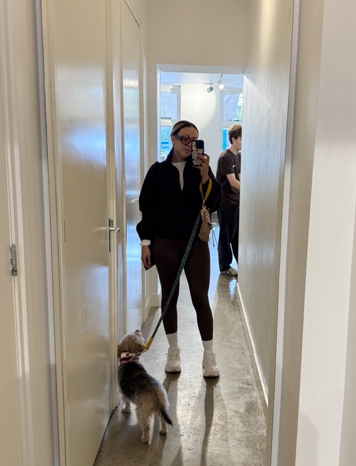 A woman takes a mirror selfie in a hallway while walking a small fluffy dog on a leash, wearing glasses, a black jacket, brown leggings, and white socks with sneakers.