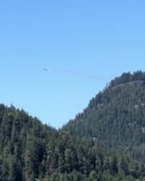 A small aircraft flies high above forested mountains, leaving a trail of white smoke against a clear blue sky.