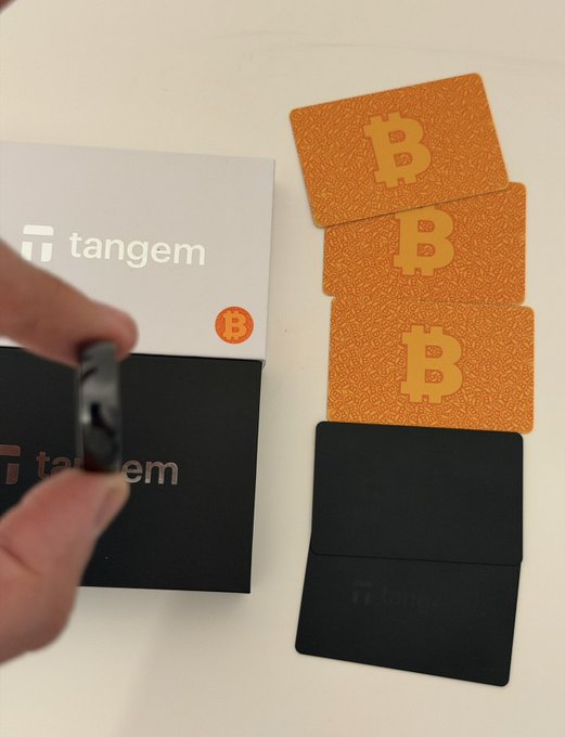 The image shows a person holding a black box with a silver logo that says "Tangem" on it. The box contains several cards, including three cards labeled "B" and one card labeled "T". These cards are likely related to Bitcoin or another digital currency. The person is standing next to the cards, possibly preparing for an event or transaction involving these cards.