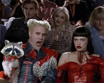 A costumed man with spiky blonde hair and a woman in a striking red corset sit in a theater audience, accompanied by a raccoon wearing a red bowtie. The image humorously captures their bold, attention-grabbing outfits, mirroring the "SO HOT RIGHT NOW" sentiment from the post.