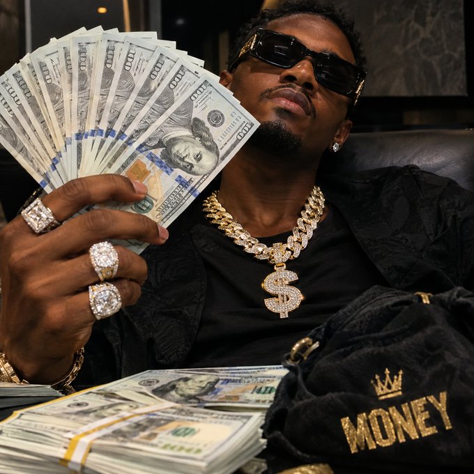 A man wearing dark sunglasses, diamond rings, and gold chains including a dollar sign pendant holds a fan of hundred-dollar bills, with more cash and a "MONEY" money bag visible.