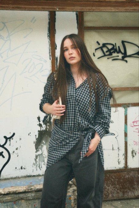 A young woman with long brown hair stands in front of a graffiti-covered wall, wearing a black and white checkered wrap shirt tied at the waist over dark pants.