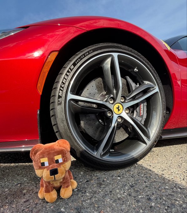 A small Minecraft-style brown bear plush sits on the pavement next to the front wheel of a red Ferrari, which features Michelin tires (size 245/35 ZR 20) and carbon ceramic brakes with the iconic prancing horse logo on the center cap—capturing a charming, playful moment perfect for the caption "Who's active?"