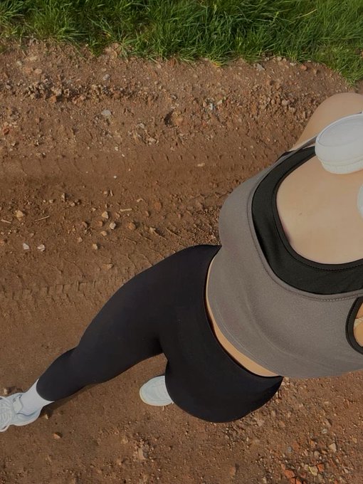 The image shows a person wearing black leggings, a gray sports bra, and white sneakers standing on dirt ground with their head tilted back as if they are about to take a selfie or share something interesting with others online. The photo appears to be taken from an elevated perspective, possibly capturing the person's upper body in profile.