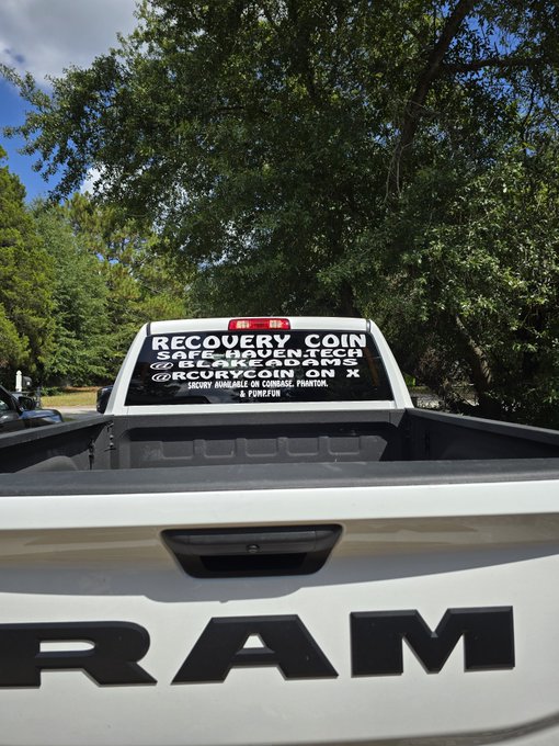 The rear window of a white RAM truck displaying text advertising 'Recovery Coin' as a safe haven, listing 'ReFi', 'GLDR', and 'ON X'.