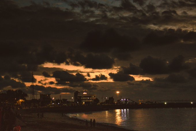 A nighttime photograph captures a beach scene with dark, dramatic clouds illuminated by an orange sunset glow on the horizon, reflecting on the water below a silhouetted cityscape.
