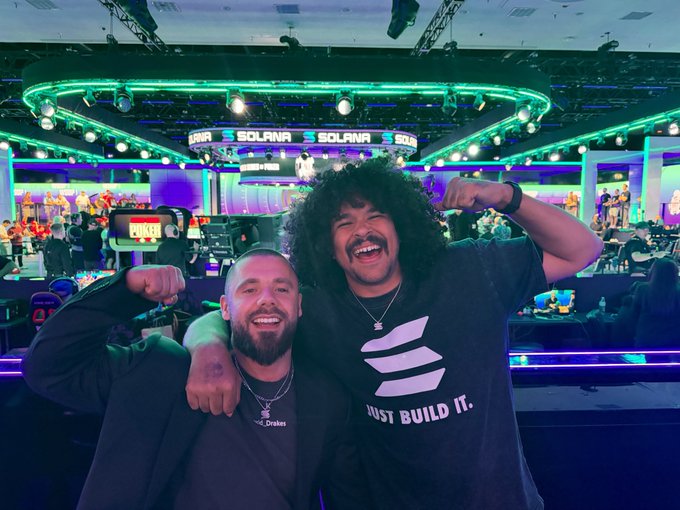 Two men pose cheerfully at what appears to be a Solana-branded event, with one wearing a black t-shirt reading 'JUST BUILD IT.' and the other wearing a 'Drake' necklace.