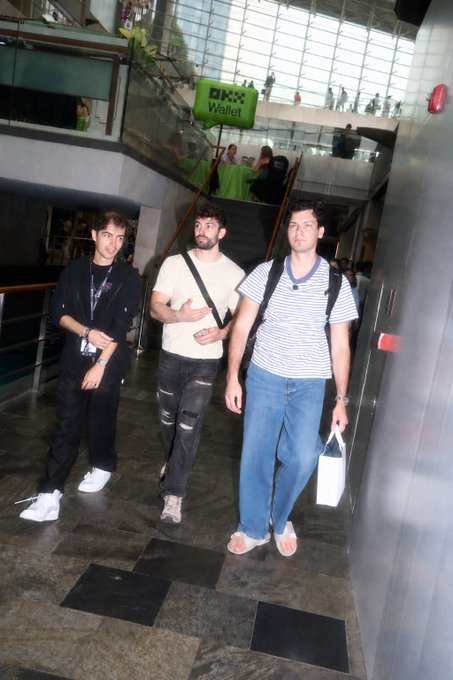 Three young men walk through a mall or convention center, with a green "OKX Wallet" sign visible in the background, suggesting the setting is a crypto/web3 conference where one of the attendees (apparently the man on the left) is being criticized for attending despite his reputation for rug-pull-related content.