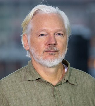 A headshot portrait of Julian Assange, the white-haired founder of WikiLeaks, wearing an olive-green button-up shirt.