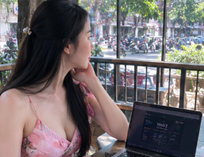 A woman with long dark hair, wearing a pink floral top, sits at a café window working on a laptop displaying an analytics dashboard, with a street scene visible behind her—visually reinforcing the post's casual morning setting while she reviews the @wallchain X Score leaderboard data on her screen.