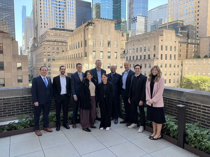 A group of nine professionally dressed individuals poses for a photo on an outdoor rooftop terrace with a city skyline and tall buildings in the background.