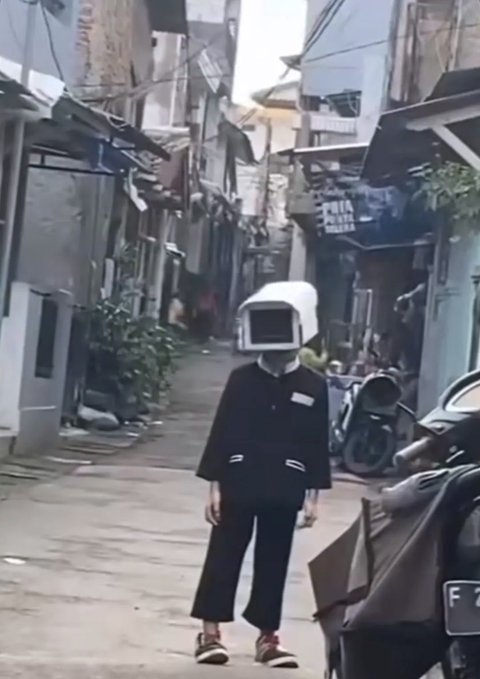 A person standing in a narrow street wearing a black outfit and an old-style computer monitor in place of a head. Parked motorcycles and buildings line the alleyway.