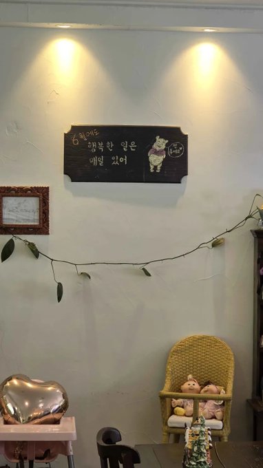 The image features a dining table with two chairs placed around it. On one side of the room, there's a black sign hanging from the wall, which reads "Happy things will happen every day." The other side of the room has a small child's chair and a pink heart-shaped balloon on the floor. A vase is also present in the scene.
