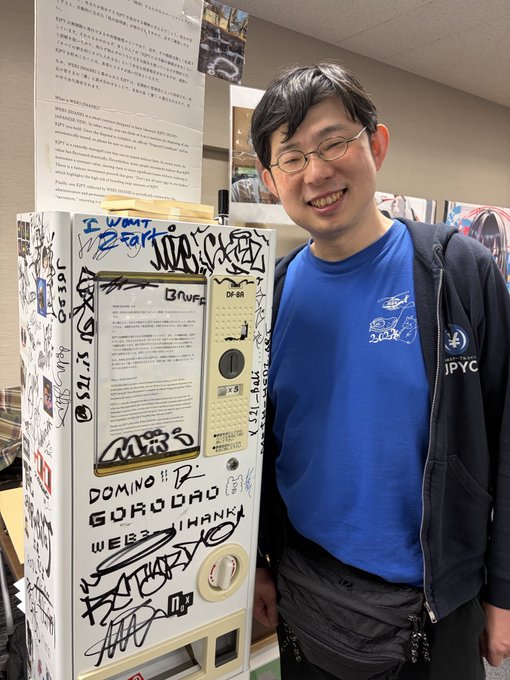 A smiling man wearing glasses and a blue t-shirt standing next to a graffiti-covered vending machine with various signatures and messages written on it.