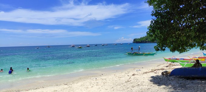 A tropical beach scene with clear turquoise water, white sand, traditional outrigger boats, and people swimming under a bright sunny sky.