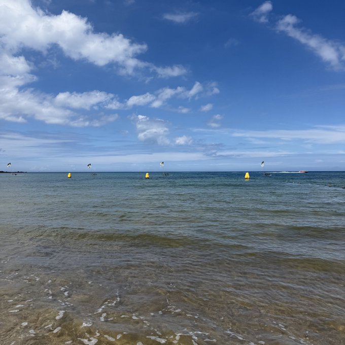 A serene coastal scene shows a clear blue sky with scattered clouds above calm ocean waters, where yellow buoys and small flags mark a swimming area in the distance.