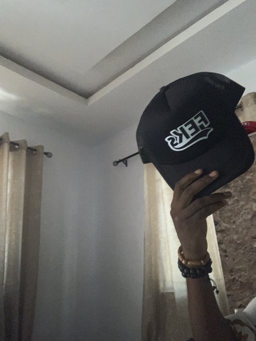 A person holding up a black snapback hat with the word 'VEE' embroidered on the front in white lettering, photographed indoors against a ceiling and curtain backdrop.