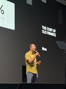 A man in a yellow shirt speaking on stage with a microphone, presenting under the title "THE COST OF OLD FINANCE."