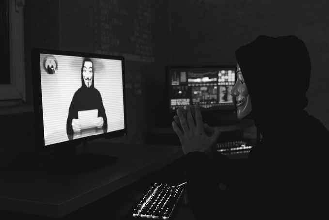 A hooded figure wearing a Guy Fawkes mask sits before multiple glowing monitors, including one displaying the Anonymous logo, casting their hands toward the screen in a dark room. The masked anonymous imagery visually reinforces the post's reference to onchain security breaches and the unidentified threat actors behind the protocols losing over $35 million.