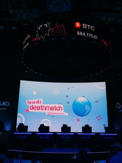 A view of the "hudi: deathmatch @basecamp" stage at Token2049, featuring five trader stations illuminated beneath a curved digital ticker displaying BTC at $84,175.01 (-1.45%) and SOL, setting the scene for the $50,000 trading competition between MartyParty, MINHxDYNASTY, Tradermayne, and CryptoKaleo.