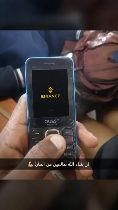 A hand holds a feature phone displaying the Binance logo, with Arabic subtitles reading "ان شاء الله طالعين من الحارة" ("God willing, we're coming out of the neighborhood"), symbolizing humble beginnings reaching toward a major global crypto platform. The image reflects the post's message of rising "from the neighborhood to the global stage."