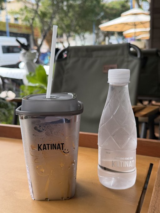 Two beverages on a wooden table at KATINAT: an iced drink in a plastic cup with straw on the left, and a bottled water with a diamond-patterned design on the right, set in an outdoor cafe.
