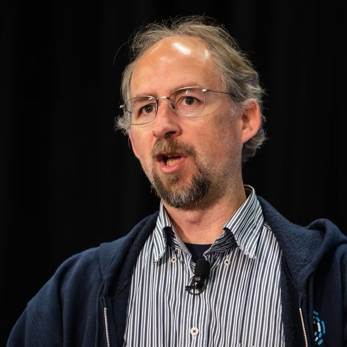 A middle-aged man with glasses, a beard, and grey hair speaks while wearing a blue hoodie over a striped shirt, with a small microphone clipped to his collar.