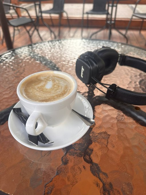 A cappuccino in a white cup sits on a saucer with a sugar packet and spoon, placed on a textured glass table beside black Bose headphones.