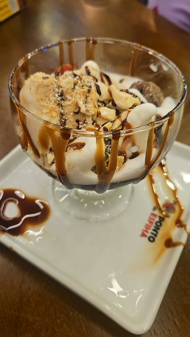 An ice cream sundae in a glass dish topped with caramel drizzle, crumbled cookies, and chocolate sauce, served on a Domino's-branded plate.